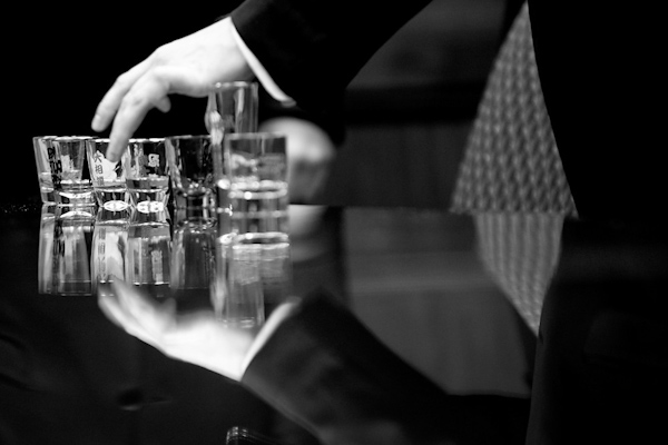 mans hands messing with shot glasses on a reflective table top- black and white - photo by Houston based wedding photographer Adam Nyholt